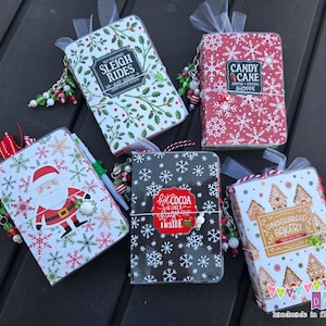 May include: Five Christmas-themed mini notebooks with decorative charms. The notebooks feature festive designs including snowflakes, Santa Claus, holly, and gingerbread houses. Each notebook has a different text label, such as "Sleigh Rides" and "Hot Cocoa".