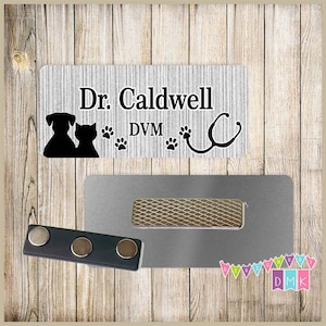 May include: A silver name tag with the text "Dr. Caldwell DVM" and a black silhouette of a dog and cat. The name tag has a silver magnetic backing.