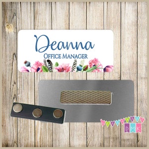 May include: White name tag with the text "Deanna Office Manager" in blue lettering. The name tag is decorated with watercolor pink flowers, blue berries, and feathers. The name tag is attached to a silver magnetic name tag holder.