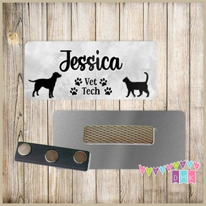 May include: A white rectangular name tag with a grey watercolour background. The name "Jessica" is written in black cursive script. Below the name is the text "Vet Tech" with paw prints on either side. A black silhouette of a dog and a cat are on either side of the text. The name tag is attached to a silver magnetic name tag holder.