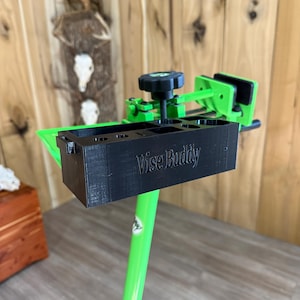 May include: A black and green vise stand with the text "Vise Buddy" on the front. The stand has a black storage compartment and a green frame. The background is a wooden wall.