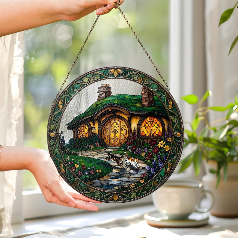K&ouml;nnte beinhalten: Ein rundes Buntglasfenster, das ein Hobbit-Haus mit gr&uuml;nem Dach, runden gelben T&uuml;ren und leuchtenden Fenstern zeigt. Ein Steinweg f&uuml;hrt zum Haus, mit bunten Blumen und zwei Katzen. Das Fenster hat einen gr&uuml;n-goldenen Rahmen und h&auml;ngt an einer Kette.