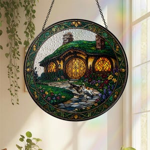 K&ouml;nnte beinhalten: Rundes Buntglasfenster, das ein Hobbit-Haus mit gr&uuml;nem Dach, gew&ouml;lbter T&uuml;r und zwei Katzen auf einem Steinweg zeigt. Das Fenster hat einen gr&uuml;nen und goldenen Rahmen und wird von einer Kette gehalten.