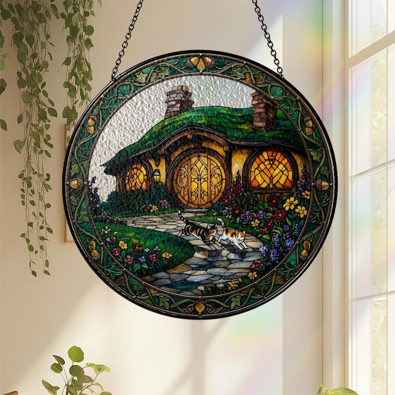 K&ouml;nnte beinhalten: Rundes Buntglasfenster, das ein Hobbit-Haus mit gr&uuml;nem Dach, gew&ouml;lbter T&uuml;r und zwei Katzen auf einem Steinweg zeigt. Das Fenster hat einen gr&uuml;nen und goldenen Rahmen und wird von einer Kette gehalten.