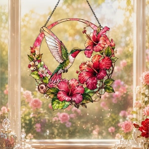 Op de afbeelding: Een ronde glas-in-lood zanger met een vliegende kolibrie omringd door felrode hibiscusbloemen en groene bladeren. Het object hangt aan een zwarte metalen ketting en toont gedetailleerde patronen in roze, groen en rood.