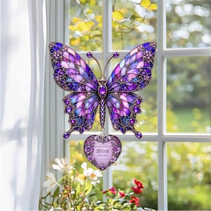 May include: A stained-glass style butterfly suncatcher in shades of purple, blue, and gold, hanging in front of a window. A heart-shaped pendant below reads "In loving memory of Mom 1969-2024". The butterfly has intricate details.