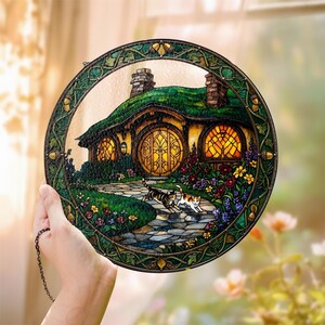K&ouml;nnte beinhalten: Runde Glasmalerei mit einem Hobbit-Haus mit gr&uuml;nem Dach, runden Fenstern und einem Steinweg. Zwei Katzen gehen den Weg entlang. Das Panel hat einen gr&uuml;nen Rahmen mit Eicheldetails.