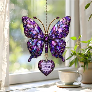 May include: A stained glass butterfly suncatcher in shades of purple, blue, and gold, suspended by a chain. A heart-shaped pendant below reads "In loving memory of Mom 1944-2026". The butterfly has intricate details and is displayed near a window.