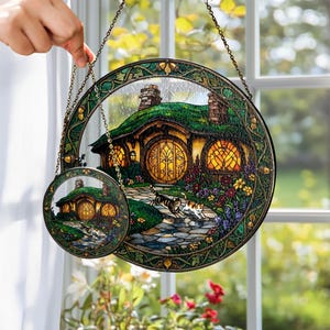 K&ouml;nnte beinhalten: Zwei runde Fensterbilder aus Glas mit einem Hobbit-Haus mit gr&uuml;nem Dach, gelben Fenstern und einem Steinweg. Das gr&ouml;&szlig;ere Bild hat einen Durchmesser von etwa 20 cm, das kleinere etwa 10 cm. Beide haben einen dekorativen Rand und eine Kette zum Aufh&auml;ngen.