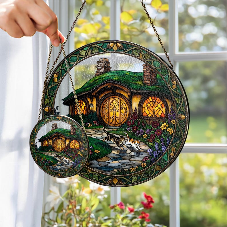 K&ouml;nnte beinhalten: Zwei runde Fensterbilder aus Glas mit einem Hobbit-Haus mit gr&uuml;nem Dach, gelben Fenstern und einem Steinweg. Das gr&ouml;&szlig;ere Bild hat einen Durchmesser von etwa 20 cm, das kleinere etwa 10 cm. Beide haben einen dekorativen Rand und eine Kette zum Aufh&auml;ngen.