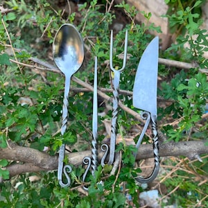 Könnte beinhalten: Ein Set aus vier handgeschmiedeten Metallutensilien, bestehend aus einem Löffel, einem dünnen Spieß, einer zweizinkigen Gabel und einem Messer. Jedes Teil hat einen gedrehten Griff, der in einer dekorativen, eingerollten Schlaufe endet.