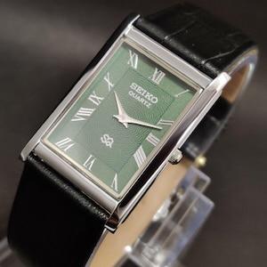 May include: A rectangular Seiko Quartz wristwatch featuring a green dial with a textured wave pattern, silver-tone Roman numeral hour markers, and silver hands. The watch has a polished silver-tone case and a black leather strap with a textured finish.