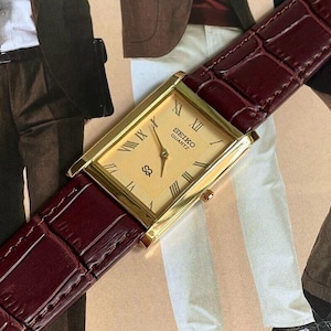 Peut inclure: Montre Seiko Quartz dorée avec un cadran rectangulaire et des chiffres romains. La montre a un cadran clair et un bracelet en cuir bordeaux avec un motif crocodile.