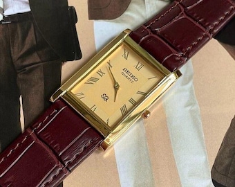 May include: A gold Seiko Quartz watch with a rectangular face and Roman numerals. The watch features a light-coloured dial and a burgundy leather strap with a crocodile pattern.