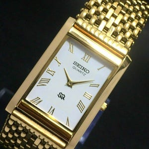 May include: A gold-toned Seiko Quartz watch with a rectangular face and a mesh band. The watch face is white with gold Roman numerals and hands. The watch has a gold bezel and band.