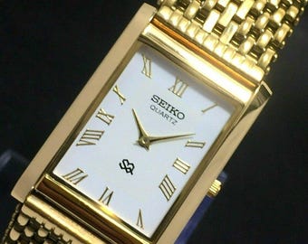May include: A gold-coloured Seiko Quartz watch with a rectangular face and a mesh strap. The watch face is white with gold Roman numerals and hands. The watch has a gold bezel and strap.