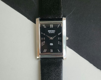 May include: A rectangular Seiko Quartz wristwatch featuring a black dial with silver-toned Roman numeral hour markers, silver hands, and a black leather strap. The watch face displays the Seiko Quartz logo and a small square emblem at the bottom.