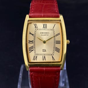 Op de afbeelding: Een goudkleurig Seiko Quartz horloge met een rechthoekige wijzerplaat en een rode leren band. De wijzerplaat van het horloge heeft zwarte Romeinse cijfers, zwarte lijnen en de woorden "SEIKO QUARTZ". Het horloge heeft een goudkleurige kroon en een rode leren band.