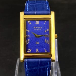 May include: A gold-toned rectangular Seiko Quartz watch with a blue face and blue leather band. The watch face features gold Roman numerals and hands. The band is a textured blue leather.