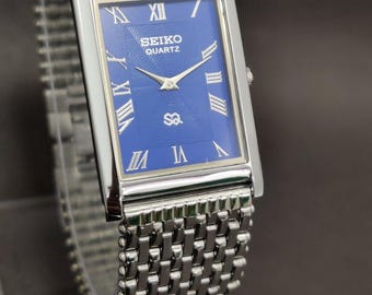 May include: A rectangular Seiko Quartz wristwatch featuring a deep blue dial with silver-toned Roman numeral hour markers and slender hands. The watch has a polished silver-tone metal case and a matching multi-link metal bracelet strap.