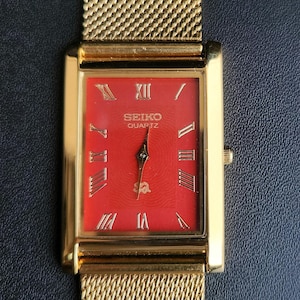 May include: A rectangular Seiko Quartz wristwatch featuring a vibrant red dial with gold-tone Roman numeral hour markers and matching gold-tone hands. The watch has a polished gold-tone metal case and a flexible gold-tone mesh metal strap.