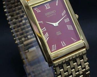 May include: A gold-plated Seiko Quartz watch with a rectangular burgundy face and Roman numerals. The watch has a gold mesh strap. The watch face displays the Seiko Quartz logo. The watch face is approximately 2.5 cm wide.