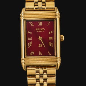 May include: A gold-tone rectangular wristwatch featuring a deep red dial with gold Roman numeral hour markers. The watch has a matching gold-tone metal link bracelet, a crown on the right side, and the text SEIKO QUARTZ printed on the dial.