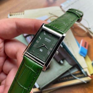 Könnte beinhalten: Eine rechteckige Seiko Quartz Armbanduhr mit einem dunkelgrünen Zifferblatt mit strukturiertem Wellenmuster, silberfarbenen römischen Ziffern als Stundenmarkierungen und einem passenden grünen Lederarmband mit Krokodilprägung und silberner Schließe.