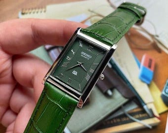May include: A rectangular Seiko Quartz wristwatch featuring a dark green dial with a textured wave pattern, silver-tone Roman numeral hour markers, and a matching green leather strap with a crocodile-embossed finish and a silver-tone buckle.