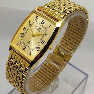 Op de afbeelding: Een goudkleurig Seiko Quartz horloge met een rechthoekige wijzerplaat en Romeinse cijfers. De band is goudkleurig en heeft een geweven stijl. De wijzerplaat toont het Seiko Quartz-logo. Het horloge wordt gepresenteerd op een transparante standaard.