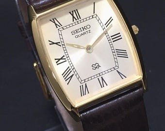 May include: A gold-coloured Seiko Quartz watch with a rectangular face and a brown leather strap. The watch face displays black Roman numerals and the Seiko Quartz logo. The watch has a classic, elegant design.