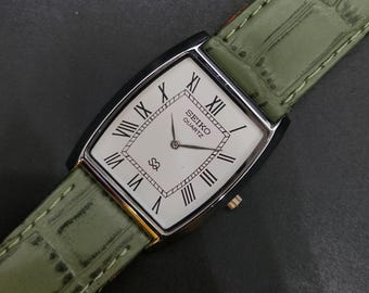 May include: A rectangular Seiko Quartz wristwatch featuring a white dial with black Roman numeral hour markers, thin black hands, and a textured sage green leather strap with a crocodile-style embossed pattern and matching stitching.