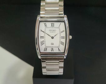 May include: A silver-toned Seiko Quartz wristwatch featuring a rectangular case and a matching link bracelet. The white dial displays black Roman numeral hour markers, thin black hands, and the Seiko Quartz logo with an SQ symbol at the bottom.