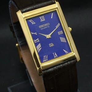 May include: A gold-coloured rectangular Seiko Quartz watch with a deep blue face and Roman numeral hour markers. The watch has gold hands and a brown leather strap.