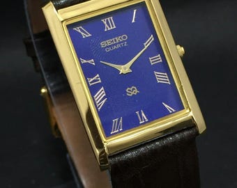 May include: A gold-coloured rectangular Seiko Quartz watch with a deep blue face and Roman numeral hour markers. The watch has gold hands and a brown leather strap.