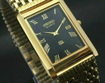 May include: A gold-coloured Seiko Quartz watch with a rectangular black face and gold Roman numerals. The watch features a gold woven strap and a small gold crown. The watch face displays the Seiko Quartz logo.