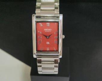 May include: A rectangular Seiko Quartz wristwatch featuring a vibrant red dial with silver-toned Roman numeral hour markers. The watch has a polished silver-toned stainless steel case and a matching link bracelet with a secure deployment clasp.