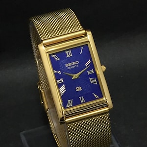 Może przedstawiać: Złoty, prostokątny zegarek Seiko Quartz z bransoletą typu mesh. Tarcza zegarka jest granatowa ze złotymi rzymskimi cyframi i wskazówkami. Zegarek ma klasyczny, elegancki design.