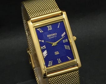 May include: Gold-toned rectangular Seiko Quartz watch with a mesh strap. The watch face is a deep blue with gold Roman numerals and hands. The watch has a classic, elegant design.