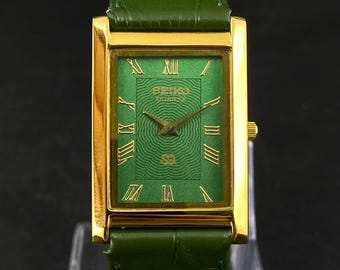 May include: A rectangular gold-toned Seiko watch with a green face and green leather strap. The watch face features Roman numerals and the Seiko logo. The strap is approximately 20 cm long.