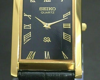 May include: A rectangular gold-toned Seiko Quartz watch with a black face and gold Roman numerals. The watch has a black leather strap. The watch face displays the Seiko logo and the word "QUARTZ".
