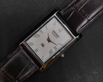 May include: A rectangular Seiko Quartz wristwatch featuring a white dial with black Roman numeral hour markers, black hands, and a logo. The watch is attached to a dark brown leather strap with a textured, embossed crocodile-style pattern.