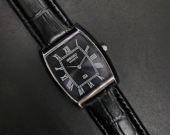 May include: A rectangular Seiko Quartz wristwatch featuring a black dial with silver-toned Roman numeral hour markers, a silver-toned case, and a black leather strap with a textured crocodile-embossed pattern and a silver-toned buckle.