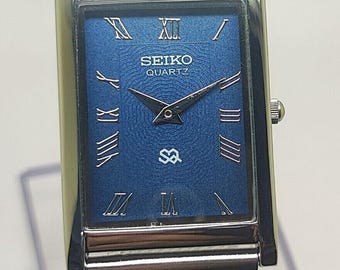 May include: A rectangular Seiko Quartz wristwatch with a textured blue dial, silver-tone Roman numeral hour markers, and silver hands. The watch features a silver-tone metal mesh strap and a polished silver-tone rectangular case with a side crown.