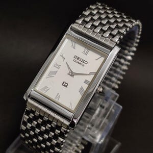 May include: A rectangular Seiko Quartz wristwatch with a silver-tone metal case and a matching multi-link bracelet. The white dial features black Roman numeral hour markers, silver hands, and the text Seiko Quartz printed in black.