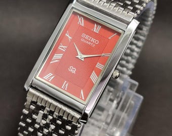 May include: A rectangular Seiko Quartz wristwatch featuring a vibrant red dial with a subtle wave pattern, silver-tone Roman numeral hour markers, and silver hands. The watch has a polished silver-tone metal case and a matching woven metal link bracelet.
