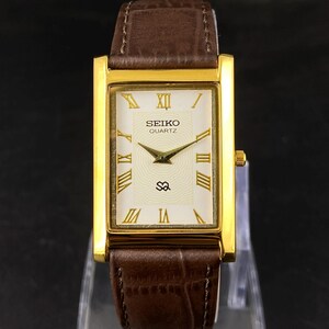 May include: A rectangular gold-toned Seiko Quartz watch with a brown leather band. The watch face is cream-colored with gold Roman numerals and hands. The band is a dark brown with a textured pattern.