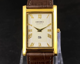 May include: A rectangular gold-toned Seiko Quartz watch with a brown leather strap. The watch face is cream-coloured with gold Roman numerals and hands. The strap is a dark brown with a textured pattern.
