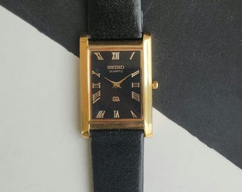 May include: A rectangular gold-toned Seiko Quartz watch with a black face and Roman numeral hour markers. The watch has a black leather strap. The watch face displays the Seiko Quartz logo and a small emblem. The watch is a classic design.