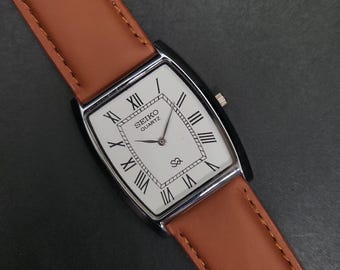 May include: A rectangular Seiko Quartz wristwatch featuring a white dial with black Roman numeral hour markers, a silver-tone case, and a tan leather strap with visible stitching. The dial includes the Seiko Quartz logo and a small SQ symbol.